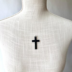 May include: A black cross pendant hangs on a mannequin. The cross is a simple, solid design, and the pendant is suspended from a thin chain. The mannequin is covered in a textured, off-white fabric.