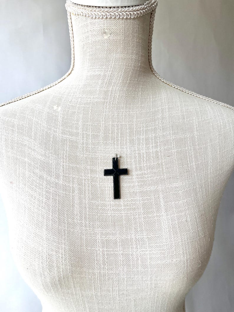 May include: A black cross pendant hangs on a mannequin. The cross is a simple, solid design, and the pendant is suspended from a thin chain. The mannequin is covered in a textured, off-white fabric.