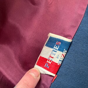 May include: Close-up of a garment's interior, showcasing a label that reads "PETITES by SUZY" in red, white, and blue. The label is sewn into a dark burgundy lining, with a navy blue fabric visible on the right side.