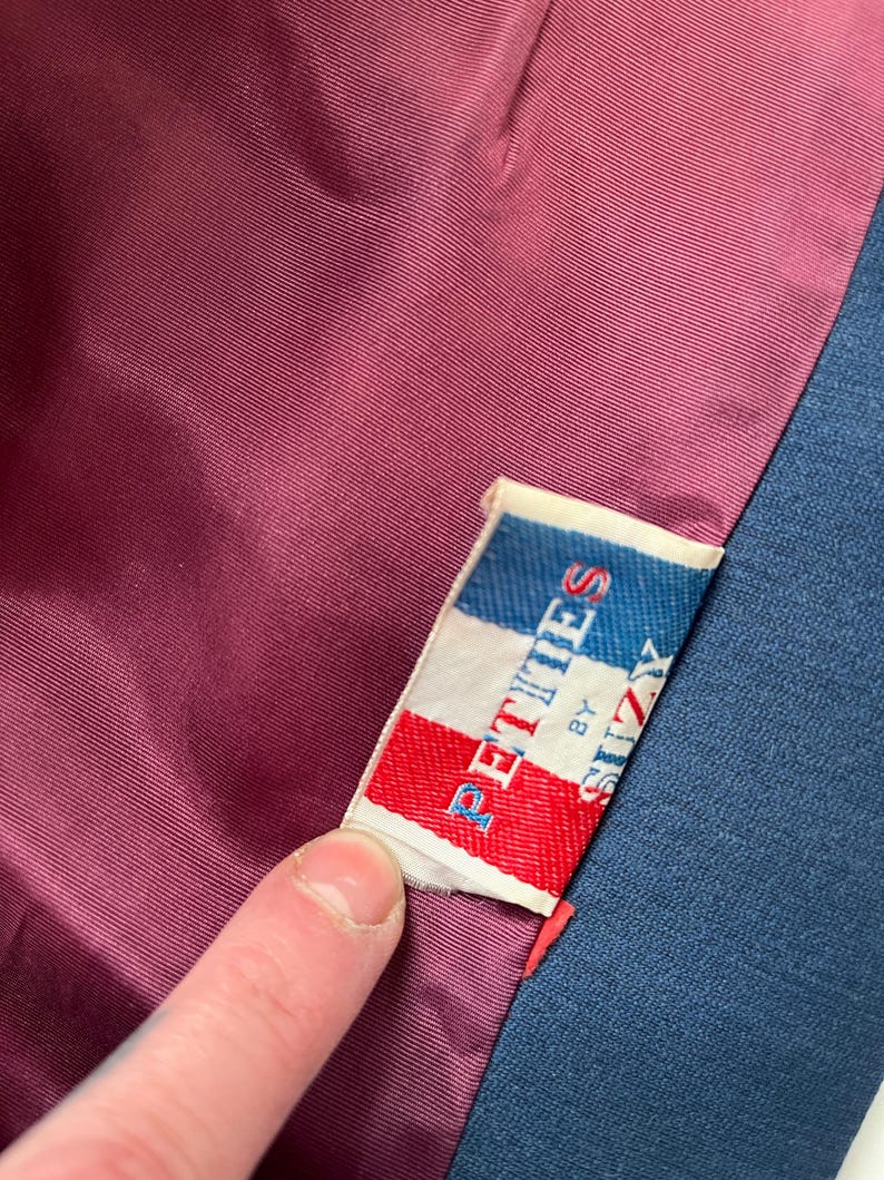 May include: Close-up of a garment's interior, showcasing a label that reads "PETITES by SUZY" in red, white, and blue. The label is sewn into a dark burgundy lining, with a navy blue fabric visible on the right side.