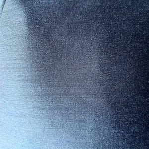 May include: Close-up of a textured, navy blue fabric. The material has a subtle sheen, with visible woven threads creating a slightly uneven surface. The color shifts slightly in the light, adding depth to the solid color.
