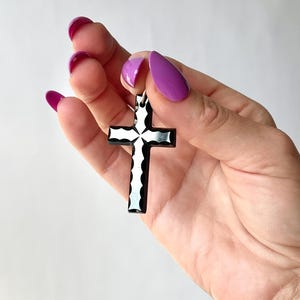May include: A black and white cross pendant with a decorative design. The cross is held against a white background. The pendant is approximately 2 inches in length.