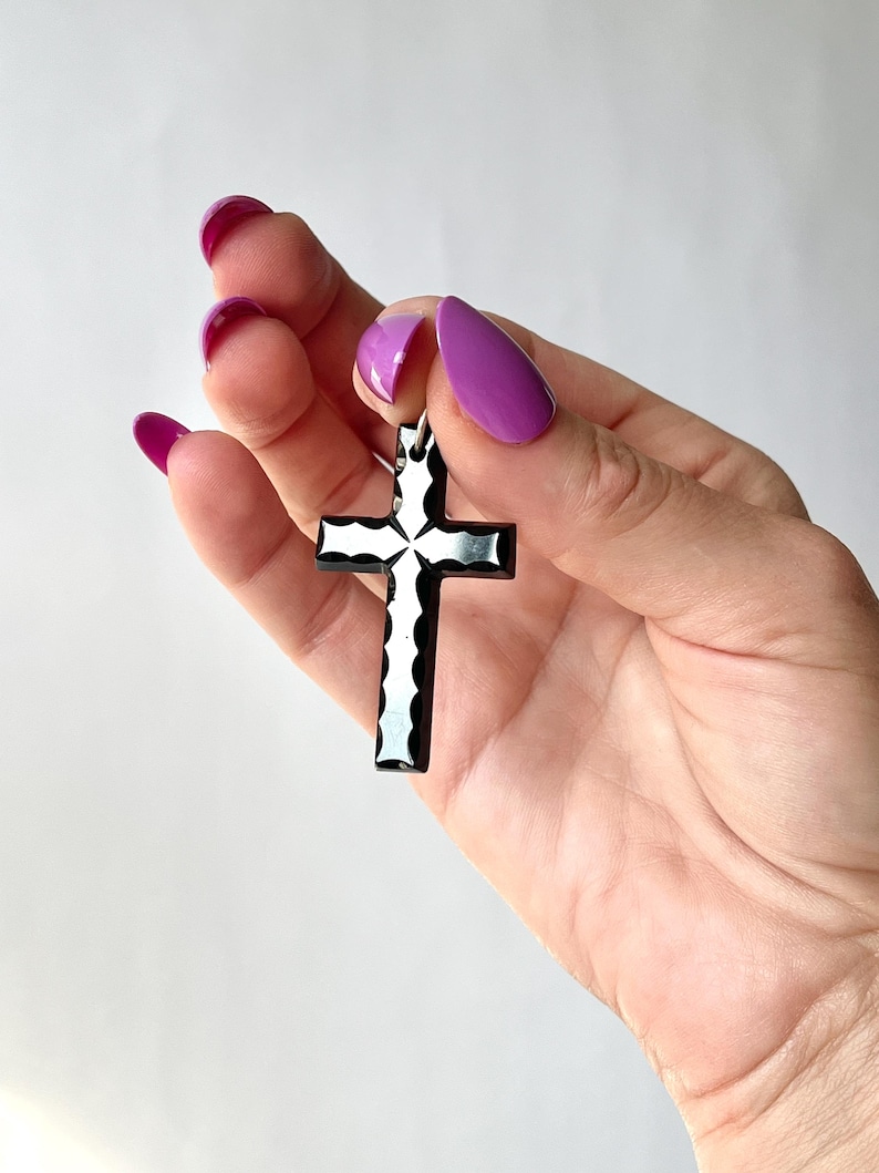 May include: A black and white cross pendant with a decorative design. The cross is held against a white background. The pendant is approximately 2 inches in length.