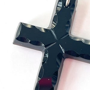 May include: A close-up of a black cross pendant with a silver-colored bail. The cross has a faceted design with a cross-shaped pattern in the center. The pendant is set against a white background.