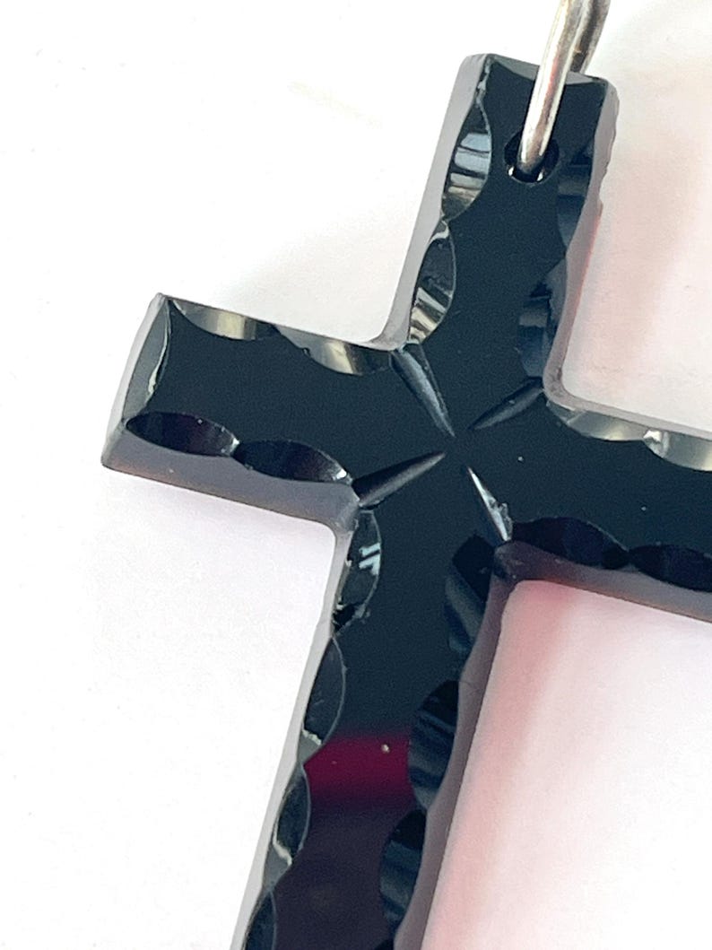 May include: A close-up of a black cross pendant with a silver-colored bail. The cross has a faceted design with a cross-shaped pattern in the center. The pendant is set against a white background.