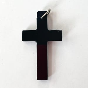 May include: A black cross pendant with a silver-colored loop at the top. The cross has a two-tone design, with the top portion black and the bottom portion a deep burgundy color. The cross is a religious symbol.