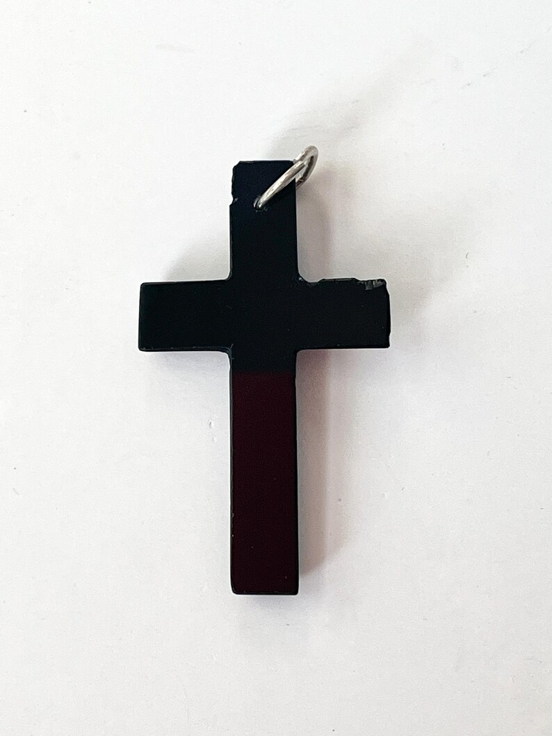 May include: A black cross pendant with a silver-colored loop at the top. The cross has a two-tone design, with the top portion black and the bottom portion a deep burgundy color. The cross is a religious symbol.
