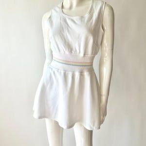 May include: White sleeveless dress with a scoop neckline and a flared skirt. The waistline features a horizontal band with pastel stripes in yellow, pink, and blue. The dress is displayed on a mannequin.