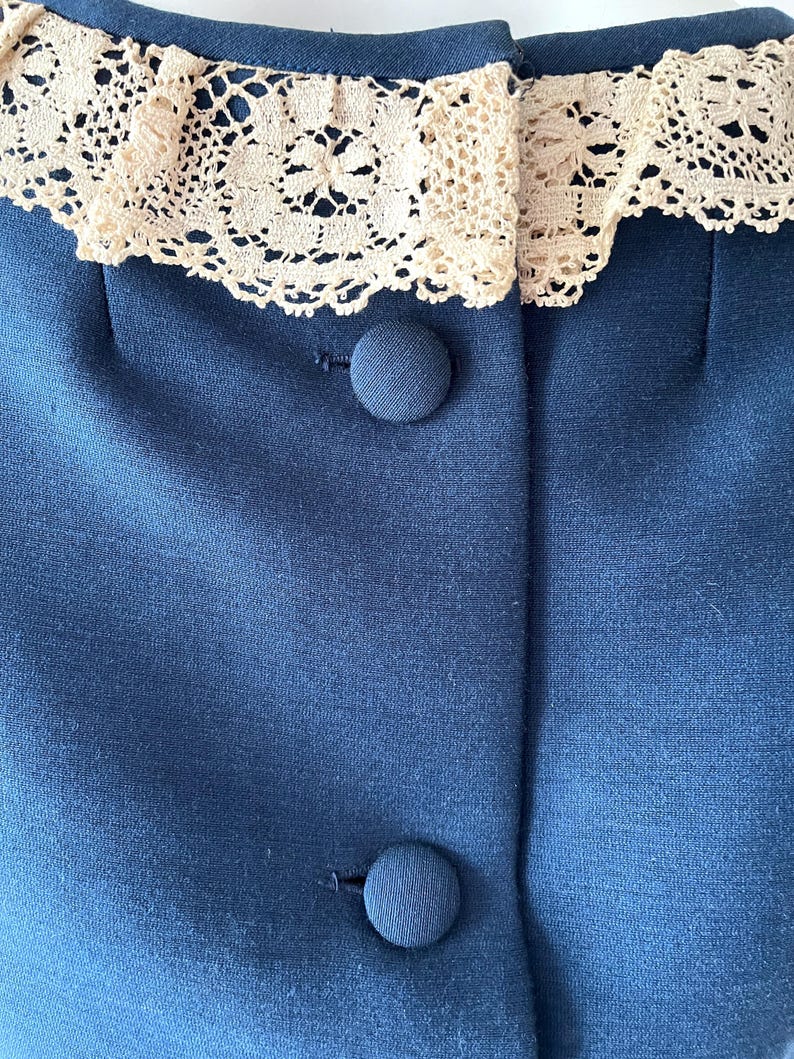 May include: Close-up of a navy blue garment with a cream-colored lace collar. The garment has two navy blue buttons. The fabric appears to be a smooth, woven material.