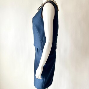 May include: A sleeveless, navy blue dress with a decorative collar. The dress has a fitted bodice and a knee-length skirt. The collar features a cream-colored lace trim and embellishments. The dress is displayed on a mannequin against a white background.