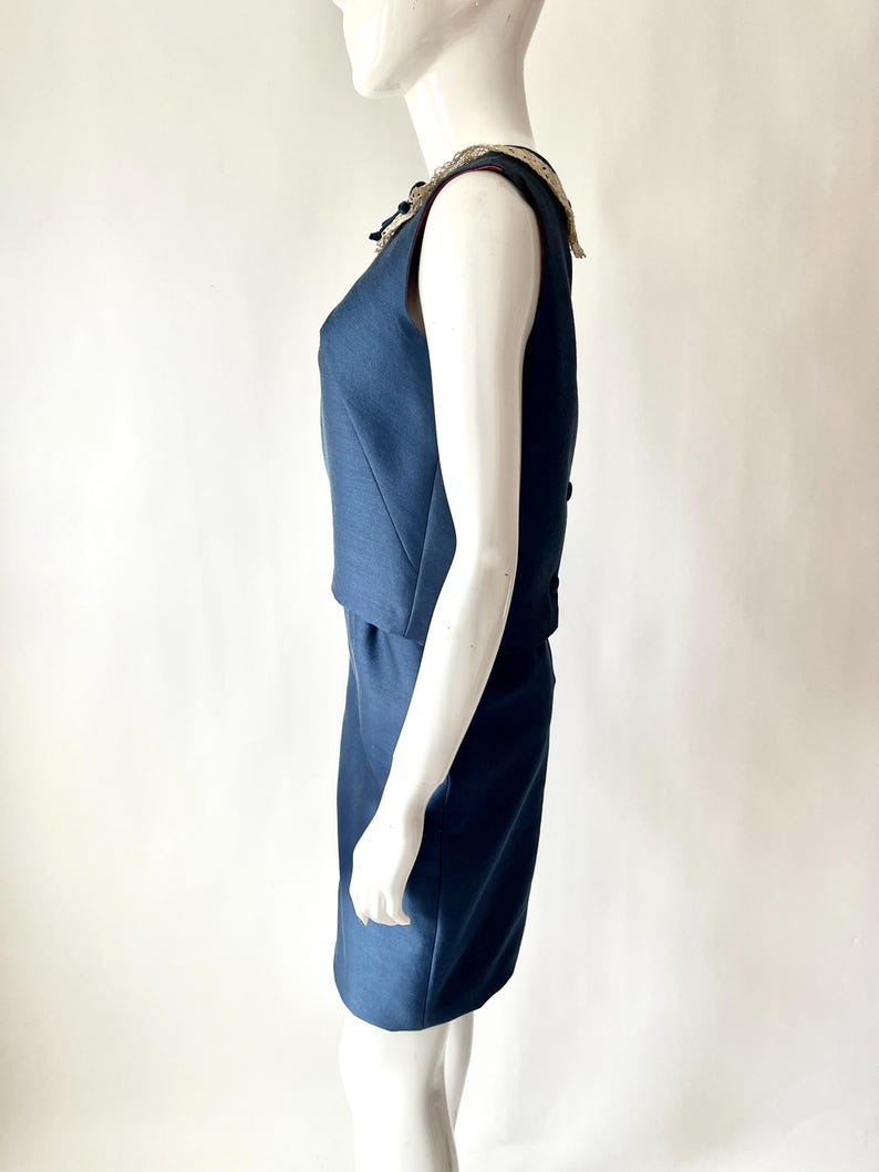 May include: A sleeveless, navy blue dress with a decorative collar. The dress has a fitted bodice and a knee-length skirt. The collar features a cream-colored lace trim and embellishments. The dress is displayed on a mannequin against a white background.