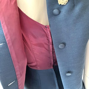 May include: A vintage blue dress with a matching vest. The vest features three round buttons and a cream-colored lace detail at the neckline. The dress has a burgundy lining and a zipper closure.