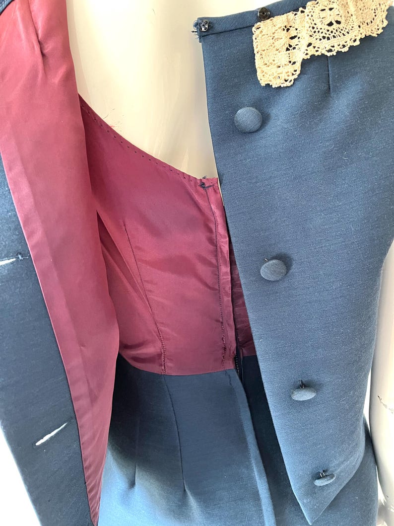 May include: A vintage blue dress with a matching vest. The vest features three round buttons and a cream-colored lace detail at the neckline. The dress has a burgundy lining and a zipper closure.