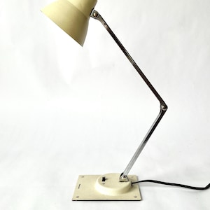 May include: Cream-colored desk lamp with a metal arm and base. The lamp features a conical shade and a square base with a circular switch. The adjustable arm allows for directional lighting. A black power cord extends from the base.