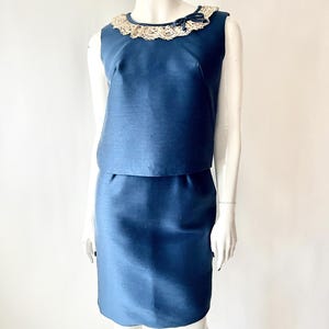 May include: A two-piece vintage outfit in a deep blue hue. The sleeveless top features a cream-colored lace collar with a small bow. The matching skirt is knee-length. The outfit is displayed on a mannequin against a white background.