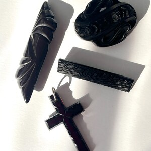May include: An assortment of black vintage jewelry pieces, including a cross pendant, a floral brooch, a barrette, and a carved art deco style pin. The items are displayed on a white surface, with shadows adding depth.