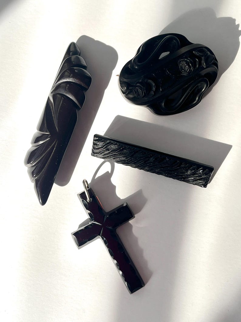 May include: An assortment of black vintage jewelry pieces, including a cross pendant, a floral brooch, a barrette, and a carved art deco style pin. The items are displayed on a white surface, with shadows adding depth.