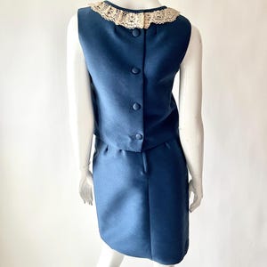 May include: A vintage navy blue two-piece dress set. The sleeveless top has a cream-colored lace collar and buttons down the back. The matching skirt is knee-length. The dress is displayed on a mannequin.