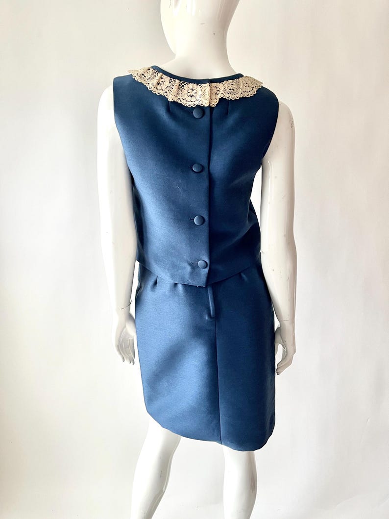 May include: A vintage navy blue two-piece dress set. The sleeveless top has a cream-colored lace collar and buttons down the back. The matching skirt is knee-length. The dress is displayed on a mannequin.