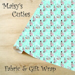Op de afbeelding: Een rol stof of cadeaupapier met een herhalend patroon van speelgoedsoldaten, schommelpaarden en ingepakte cadeaus op een lichtblauwe achtergrond. De woorden "Maisy's Cuties" en "Fabric & Gift Wrap" zijn zichtbaar.