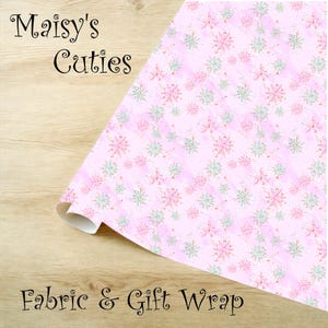 Op de afbeelding: Roze stof en cadeaupapier met een sneeuwvlokpatroon. De sneeuwvlokken zijn in roze, mintgroene en gouden tinten. De woorden "Maisy's Cuties" en "Fabric & Gift Wrap" staan op de stof.