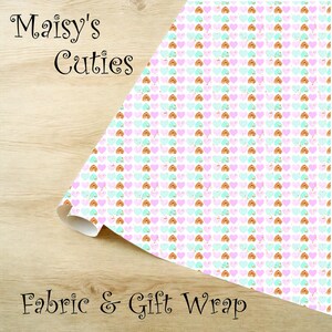 Op de afbeelding: Een rol stof of cadeaupapier met een herhalend patroon van harten in pastelkleuren: roze, mintgroen en goud. De woorden "Maisy's Cuties" en "Fabric & Gift Wrap" staan op de afbeelding.