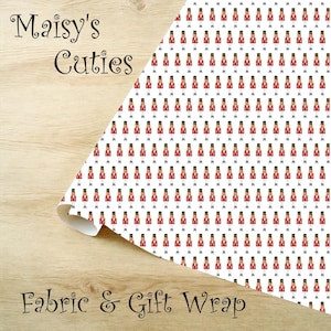 Op de afbeelding: Witte stof of cadeaupapier met een herhalend patroon van speelgoedsoldaten in rode jassen en zwarte hoeden. De woorden "Maisy's Cuties" en "Fabric & Gift Wrap" staan op de afbeelding.