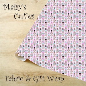 Op de afbeelding: Roze stof en cadeaupapier met een herhaald patroon van kleurrijke notenkrakerfiguren. De tekst "Maisy's Cuties" en "Fabric & Gift Wrap" zijn zichtbaar op de afbeelding.