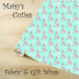 Op de afbeelding: Een rol stof en cadeaupapier met een herhalend patroon van kerstbomen. De bomen zijn wit met rode, roze en blauwe accenten, tegen een lichtblauwe achtergrond met witte stippen. De tekst "Maisy's Cuties" en "Fabric & Gift Wrap" is zichtbaar.
