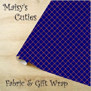 Op de afbeelding: Blauwe stof en cadeaupapier met een goudkleurig rasterpatroon. De woorden "Maisy's Cuties" en "Fabric & Gift Wrap" staan op de stof gedrukt. De stof is opgerold op een houten oppervlak.