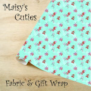 Op de afbeelding: Een rol stof en cadeaupapier met een herhalend patroon van peperkoekenmannetjes, zuurstokken en sneeuwvlokken op een lichtblauwe achtergrond. De tekst "Maisy's Cuties" en "Fabric & Gift Wrap" is afgedrukt.