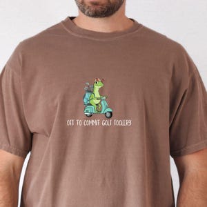 Op de afbeelding: Bruin T-shirt met een cartoon hagedis op een turquoise scooter, met golfclubs. De tekst luidt "OFF TO COMMIT GOLF FOOLERY". Het shirt heeft korte mouwen en een ronde hals. De kleur is espresso.