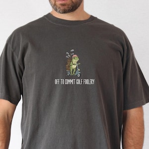 Op de afbeelding: Pepperkleurig T-shirt met een grillige afbeelding van een kikker op een golfkar met golfclubs. De tekst "OFF TO COMMIT GOLF FOOLERY" staat eronder. Het shirt heeft korte mouwen en een ronde hals.
