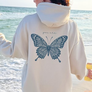 May include: A cream-colored hoodie with a blue butterfly graphic on the back. The butterfly is made up of words, including "you are" and "brave". The hoodie is worn by a person standing on a sandy beach.