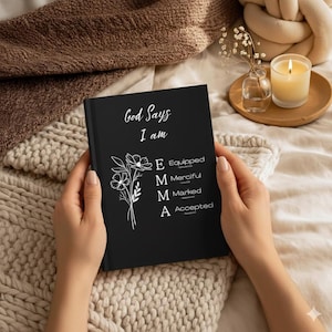 May include: A black hardcover journal featuring the white text God Says I am, a floral illustration, and the name EMMA vertically aligned with the words Equipped, Merciful, Marked, and Accepted, each accompanied by a small Bible verse reference.