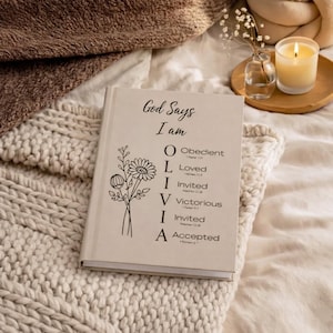 May include: A beige hardcover book with black text and a floral line drawing. The cover reads God Says I am OLIVIA, with O for Obedient, L for Loved, I for Invited, V for Victorious, I for Invited, and A for Accepted, each followed by a Bible verse.