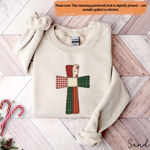 May include: A sand-colored sweatshirt featuring a digitally printed patchwork cross design. The cross is composed of various fabric patterns in red, green, and cream colors. The sweatshirt has a crew neck and long sleeves. The text "Sand" is at the bottom right.