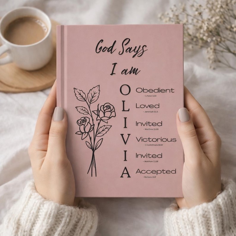 May include: A dusty pink hardcover journal featuring the black text God Says I am, a vertical acrostic for the name OLIVIA with corresponding biblical descriptors and scripture references, and a black line art illustration of a rose bouquet.