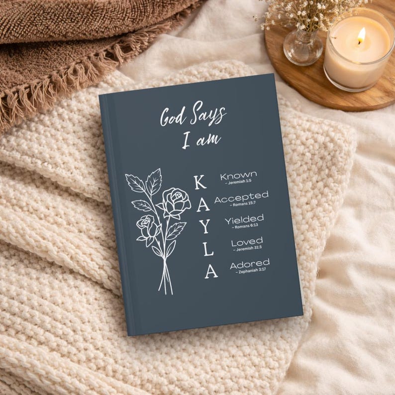 May include: A dark gray hardcover journal featuring the white text God Says I am and the name KAYLA vertically aligned with descriptive words Known, Accepted, Yielded, Loved, and Adored, accompanied by a white line drawing of a rose bouquet.