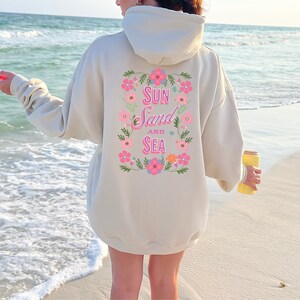 May include: A cream-colored hoodie with a floral design and the text "Sun Sand and Sea" on the back. The hoodie is being worn by a person standing on a sandy beach.