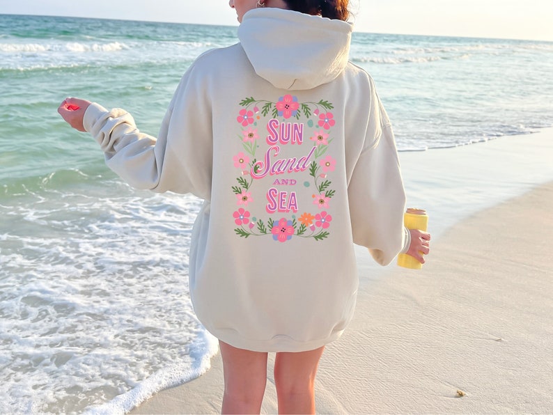 May include: A cream-colored hoodie with a floral design and the text "Sun Sand and Sea" on the back. The hoodie is being worn by a person standing on a sandy beach.