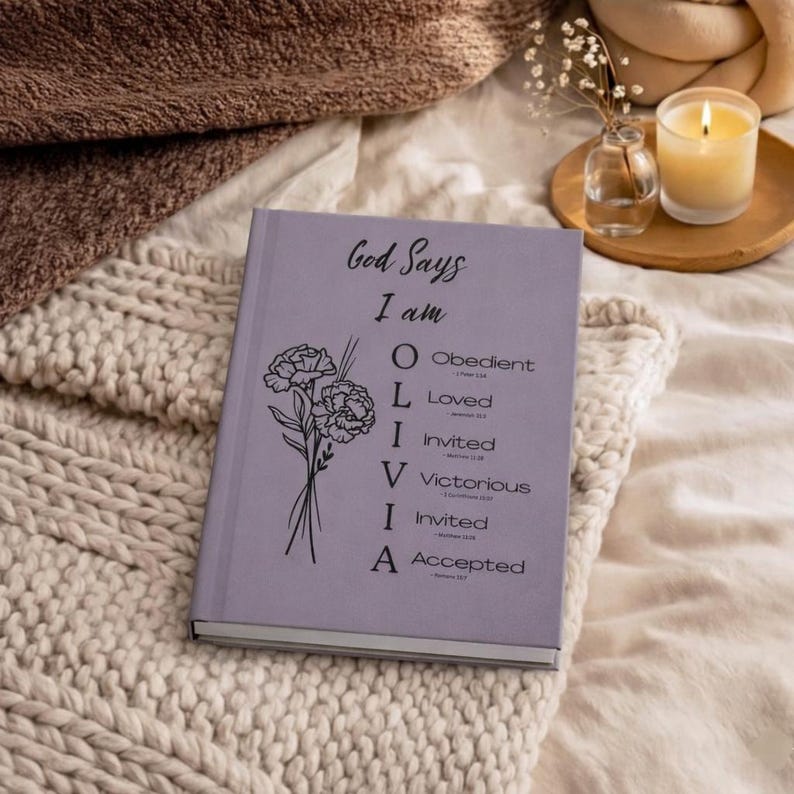 May include: A light purple hardcover book featuring a black floral illustration and the text God Says I am OLIVIA, with an acrostic list defining the name as Obedient, Loved, Invited, Victorious, Invited, and Accepted, each with a small scripture reference.