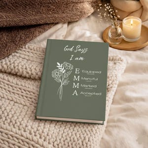 May include: A sage green hardcover journal featuring white text that reads God Says I am, followed by a floral illustration and the vertical name EMMA with the corresponding words Equipped, Merciful, Marked, and Accepted listed beside each letter.
