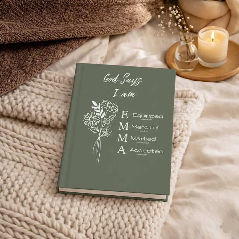 May include: A sage green hardcover journal featuring white text that reads God Says I am, followed by a floral illustration and the vertical name EMMA with the corresponding words Equipped, Merciful, Marked, and Accepted listed beside each letter.