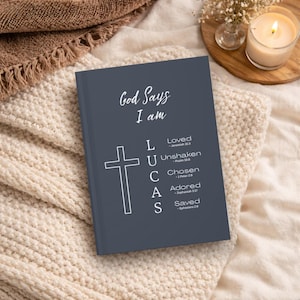 May include: A dark gray hardcover journal featuring the white text God Says I am, a cross icon, and the name LUCAS vertically aligned with the words Loved, Unshaken, Chosen, Adored, and Saved, each accompanied by a corresponding Bible verse reference.