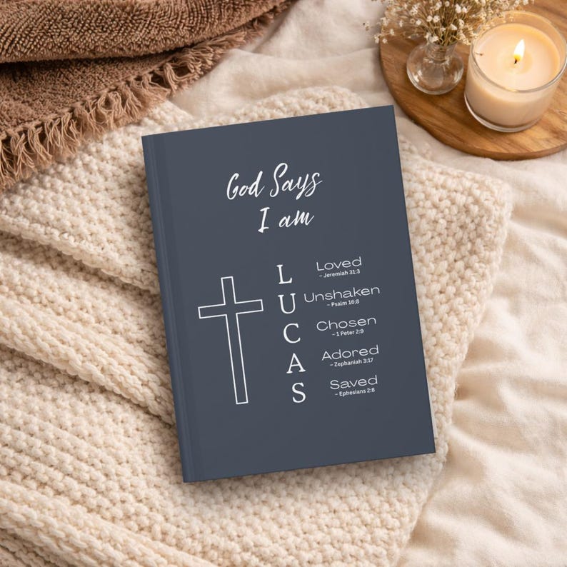 May include: A dark gray hardcover journal featuring the white text God Says I am, a cross icon, and the name LUCAS vertically aligned with the words Loved, Unshaken, Chosen, Adored, and Saved, each accompanied by a corresponding Bible verse reference.