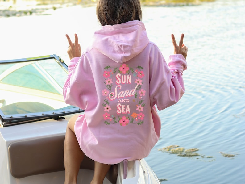 May include: Pink hoodie with a floral design and the text "Sun Sand and Sea" on the back.