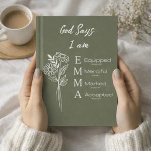 May include: A sage green journal held by hands, featuring the text "God Says I am EMMA" in white script. The journal has a floral design and the words "Equipped, Merciful, Marked, Accepted" are listed below the name.