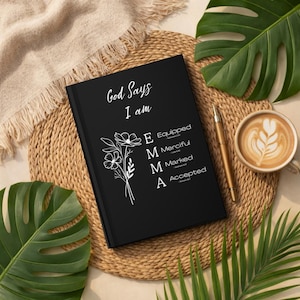 May include: A black hardcover journal featuring white text that reads God Says I am, followed by the name EMMA with each letter representing a word: Equipped, Merciful, Marked, and Accepted, accompanied by a white line-art floral illustration.