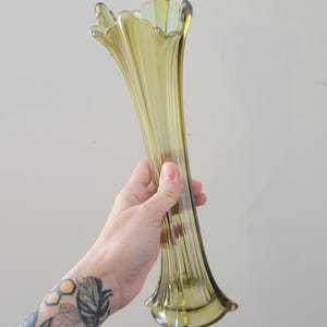 May include: A tall, green glass vase with a fluted design and a flared opening. The vase is made of iridescent glass and has a vintage look.